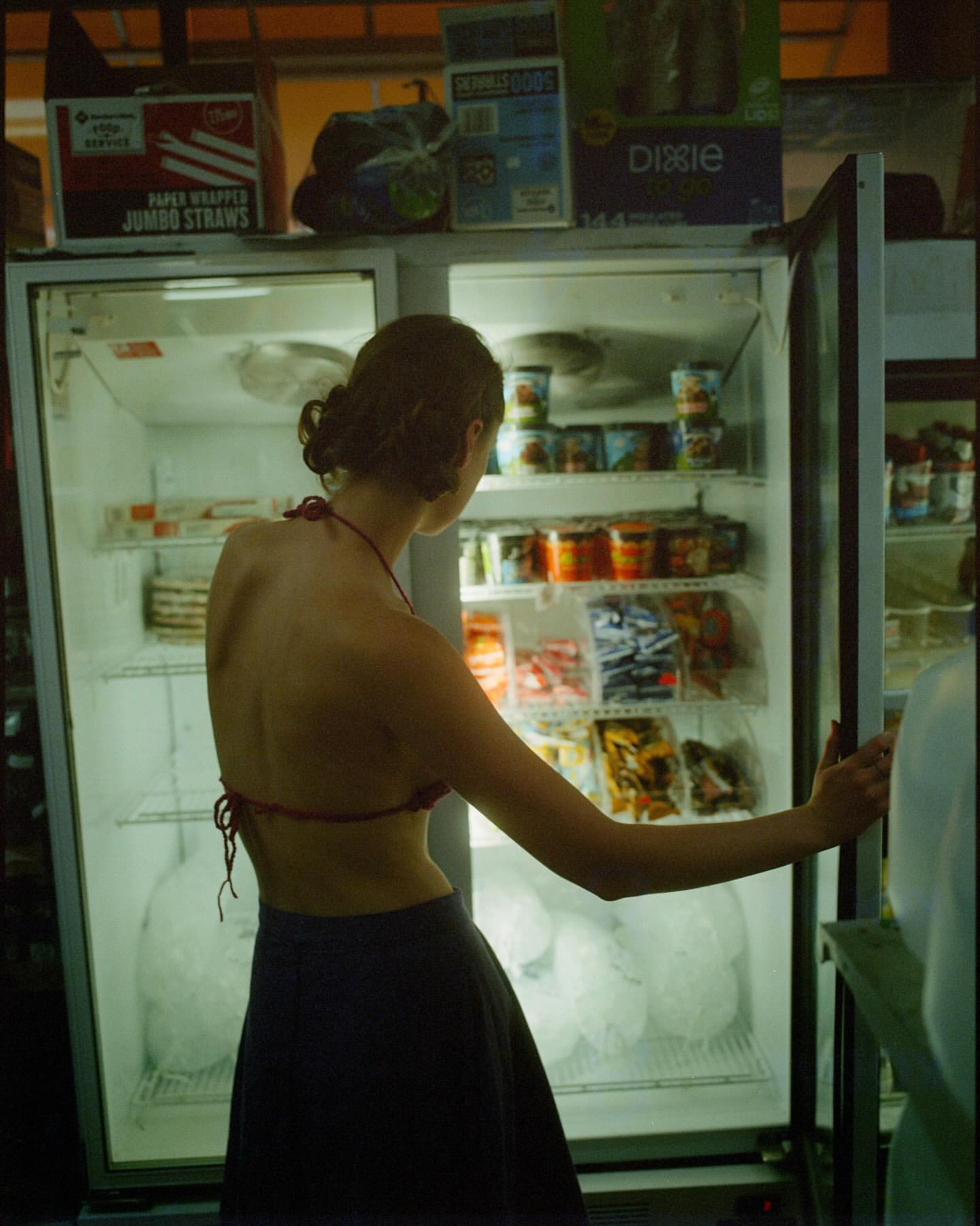 Woman opens glass door of commercial refrigerator filled with drinks and snacks in convenience store with overhead product boxes.