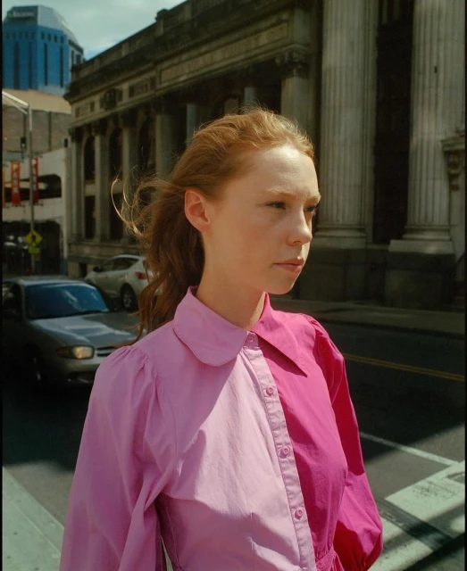 Portrait of a red-haired woman with a pink color-blocked shirt on a city street with buildings and cars in the background.