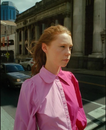 Portrait of a red-haired woman with a pink color-blocked shirt on a city street with buildings and cars in the background.