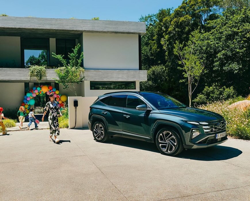 Green SUV parked in front of a modern home with a balloon arch and children. An adult woman in a dress walks towards the car.