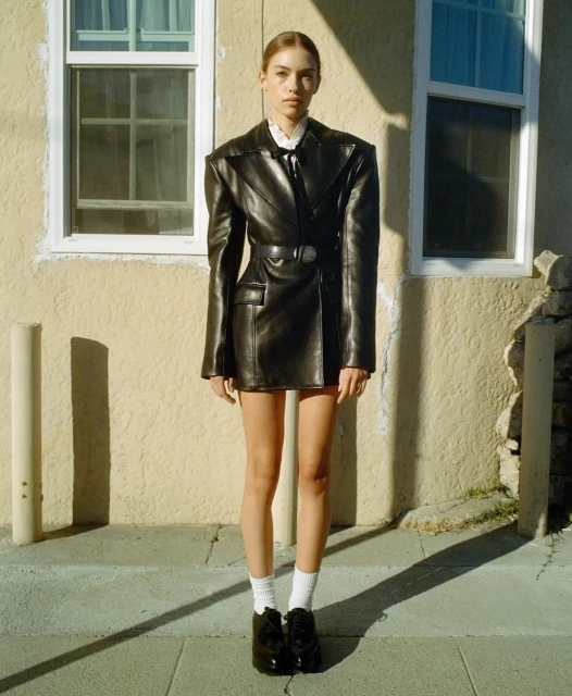 Fashion shot of a young woman in a black leather blazer dress, white shirt, and black shoes, posing outside a tan building with white trim.