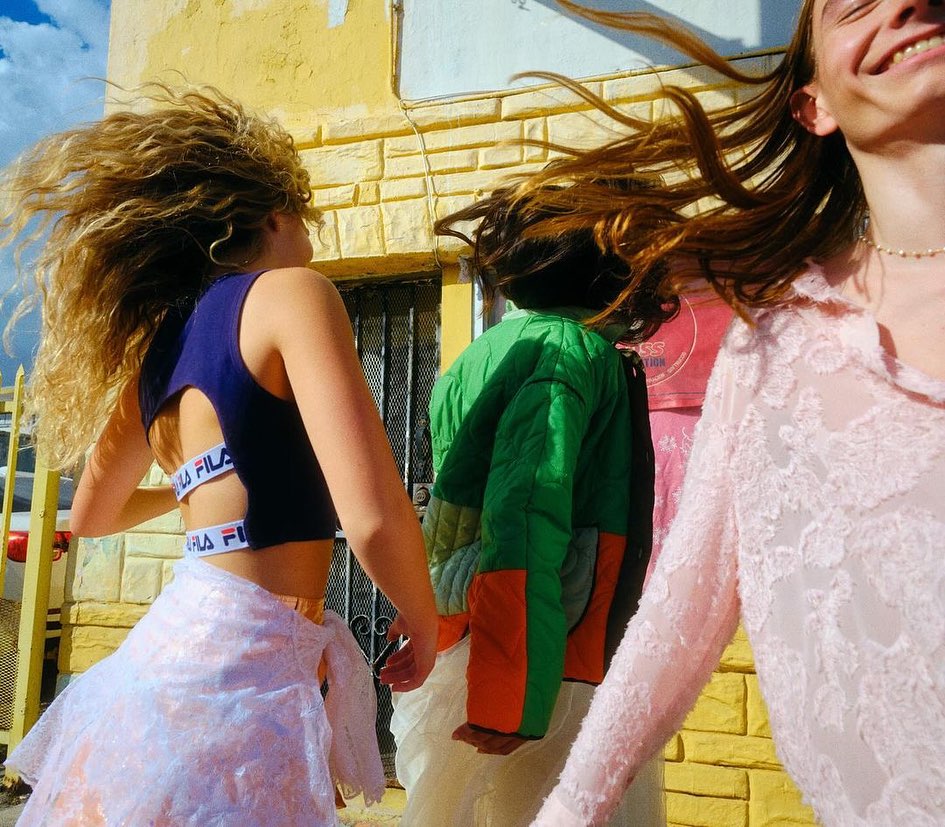 Three people with windblown hair walk near a yellow building; one wears a FILA top and lace skirt, another a color-blocked jacket.