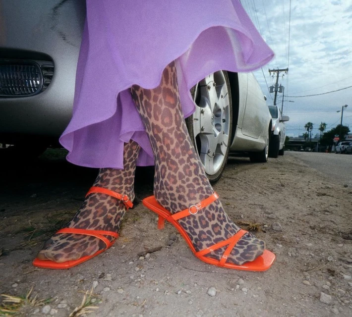 Woman in orange sandals and leopard-print tights standing next to a silver car on an outdoor street in an urban setting.