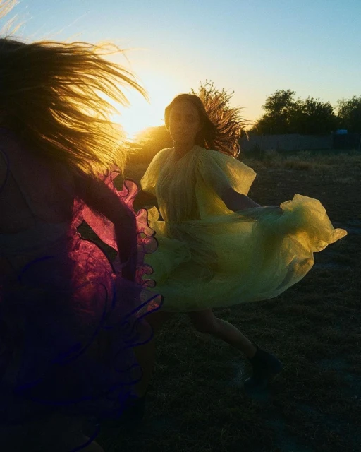 Two women in bright dresses dance in a field at sunset. The scene captures movement and the golden light of the setting sun.