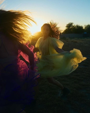 Two women in bright dresses dance in a field at sunset. The scene captures movement and the golden light of the setting sun.