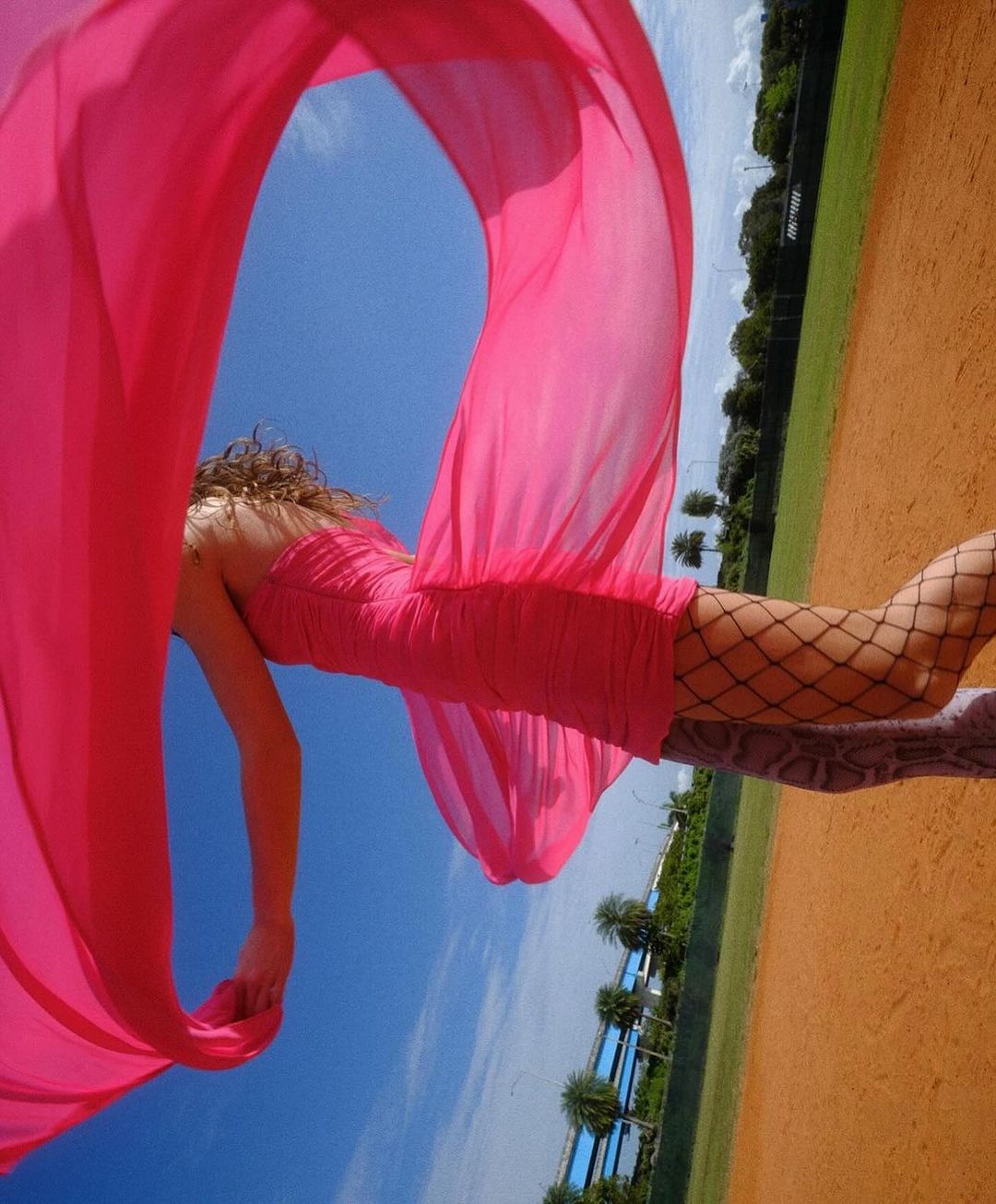 Woman in pink dress and fishnet stockings lying on baseball field with pink scarf blowing in blue sky with trees in the background.