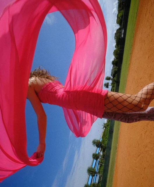 Woman in pink dress and fishnet stockings lying on baseball field with pink scarf blowing in blue sky with trees in the background.