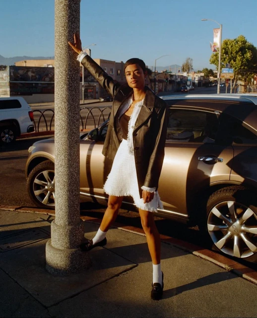 Fashionable woman poses on city street in white dress, black jacket, socks, and loafers with car in background under blue sky.