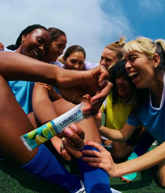 A group of smiling women in sports attire gather on a field, with one woman holding and displaying a Liquid I.V. Hydration Multiplier packet.