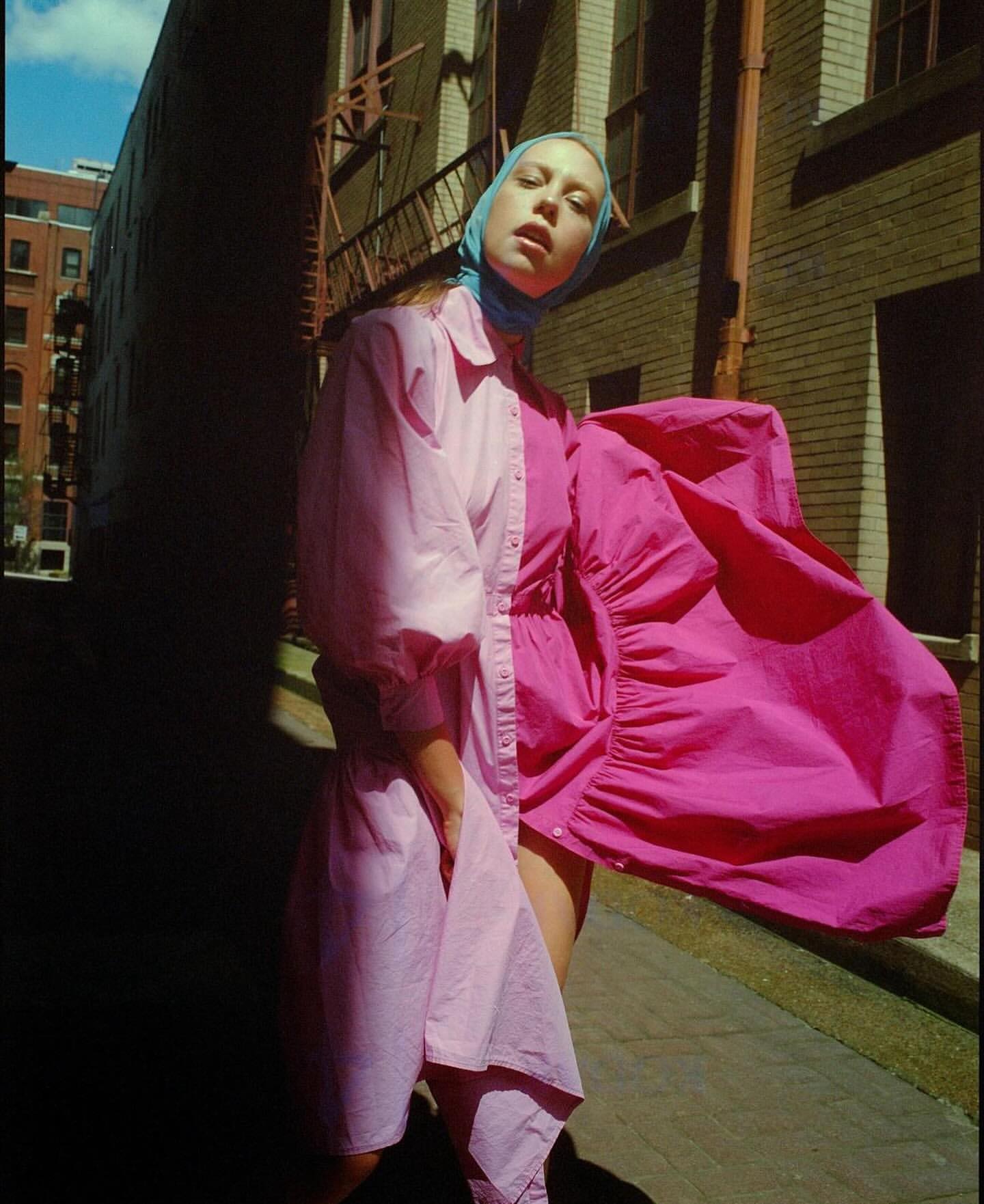 Fashion shot of a woman in a pink two-tone dress and blue headscarf, posing in an alleyway with brick buildings, capturing urban style.