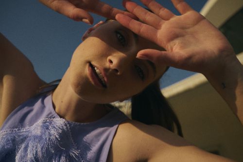 Close-up of a young woman with her hands up to shield the sun, wearing a lavender top, outdoors with a blue sky in the background.