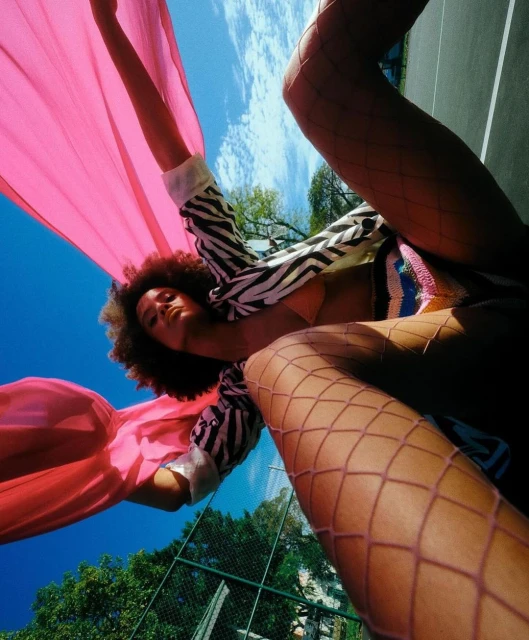 Eye-level shot of a woman with afro, wearing a zebra print jacket, pink top and fishnets, posing with a pink fabric outdoors.