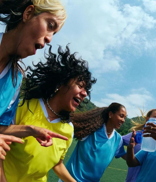 Group of smiling women in sports uniforms on a soccer field under a sunny sky, captured with a low-angle wide shot to enhance the energetic mood.