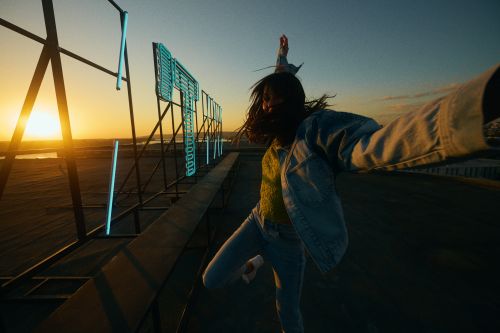 Woman in denim jacket dances near neon sign against sunset sky. Energetic pose, golden hour light, urban outdoor setting.