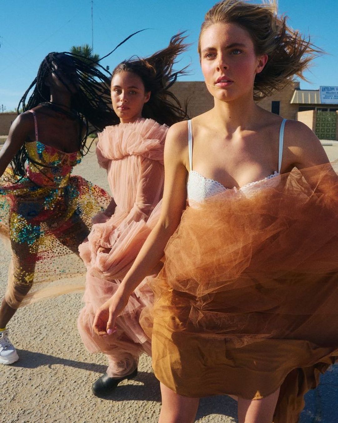 Three fashion models pose in tulle dresses outdoors, featuring diverse ethnicities and a sunlit desert environment with a building.