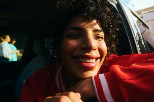 Close up of smiling woman with curly hair leaning out of a car window on a sunny day, wearing a red and white striped sweater.