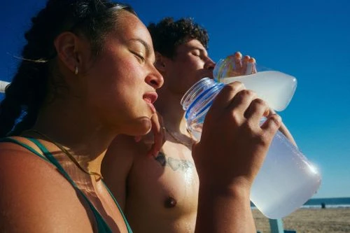Two people drinking from clear water bottles on a sunny sandy beach under a bright blue sky.