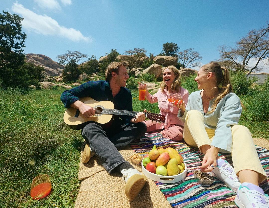 Three people enjoying a picnic outdoors on a sunny day. A man plays the guitar while two women hold drinks and smile. Trees and sky background.