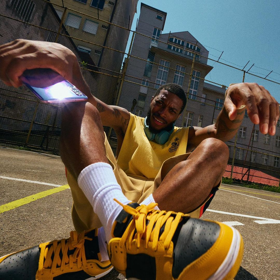 A Black male in a yellow tank top sits on a sunny outdoor court, holding a glowing smartphone with urban buildings in the background.