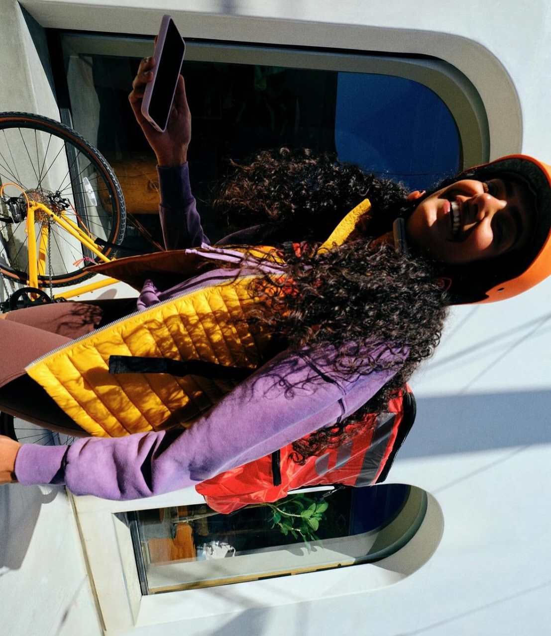 Woman in helmet smiles, holding phone near train window with a bike. She wears a vest, purple sweater and backpack.