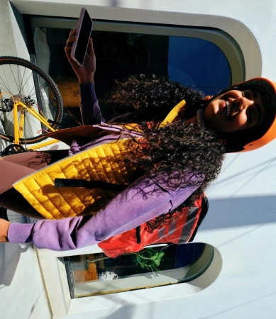 Woman in helmet smiles, holding phone near train window with a bike. She wears a vest, purple sweater and backpack.