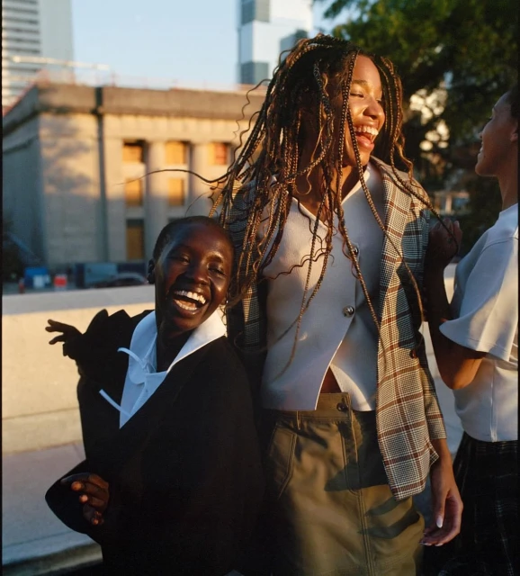 Three women laugh together outdoors in a city, wearing casual and semi-formal attire, with an urban background and golden hour lighting.