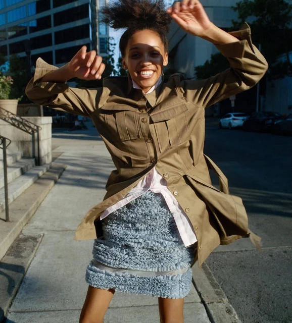 Joyful African-American woman in khaki jacket and grey ruffle skirt leaps mid-air on a sunlit city sidewalk with office buildings.