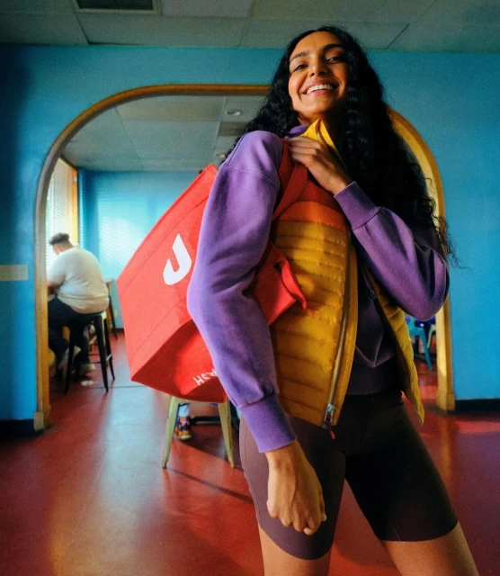 Smiling woman with a red insulated delivery bag standing indoors with a partial view of a second person at a table in the background.