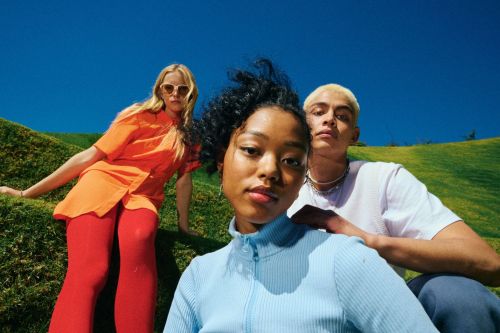 Three diverse adults pose outdoors on a grassy hill under a bright blue sky, wearing casual vibrant clothing and fashionable sunglasses.