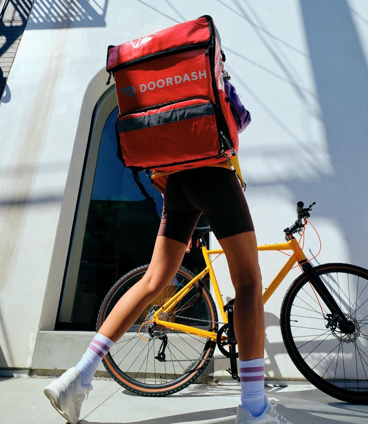 Delivery cyclist with DoorDash backpack standing next to bicycle on city street. Red backpack dominant with white building in background.