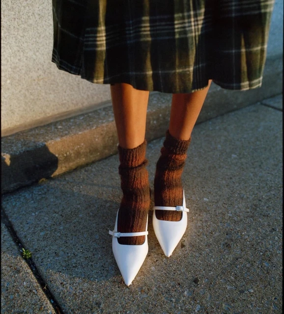 Woman stands on concrete in white Mary Jane heels, brown socks, and a green plaid skirt with diffused, warm lighting.
