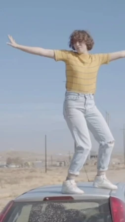 Woman stands atop a car with arms outstretched in desert landscape under blue sky, wearing jeans, yellow shirt, and white sneakers.