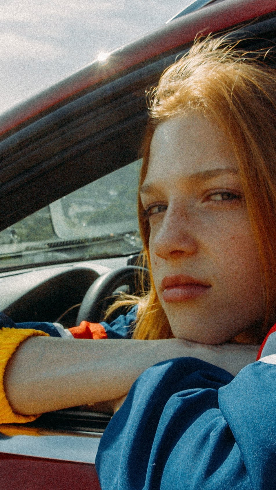 Portrait of a young redhead woman with freckles resting her head on her arm while sitting in the driver's seat of a red convertible car.