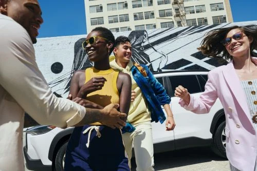 A group of young adults joyfully poses in front of a white SUV with an artistic mural on the building behind them in bright daylight.