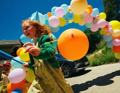 A young girl in a party hat runs outdoors with balloons at a birthday celebration with other children and a car in the background.