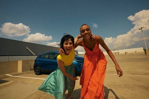Two women smiling and embracing next to a parked blue SUV against a bright sky, creating a joyful and vibrant automotive lifestyle image.