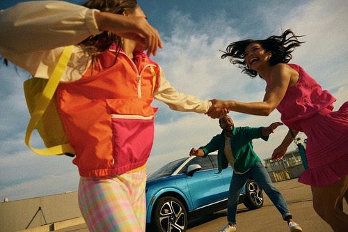 Three people are laughing and holding hands near a light-blue car under a partly cloudy sky in an outdoor urban environment. 