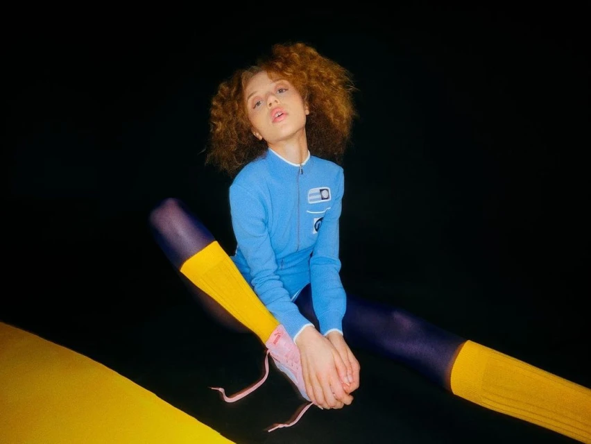 Young woman with curly hair sits in a studio wearing a blue suit, yellow socks, and pink sneakers against a black backdrop.