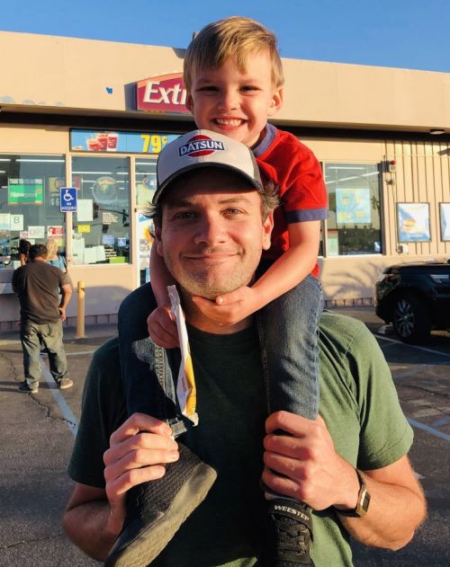 A happy child sits on an adult's shoulders in front of a convenience store with blue sky, creating a heartwarming, candid scene.