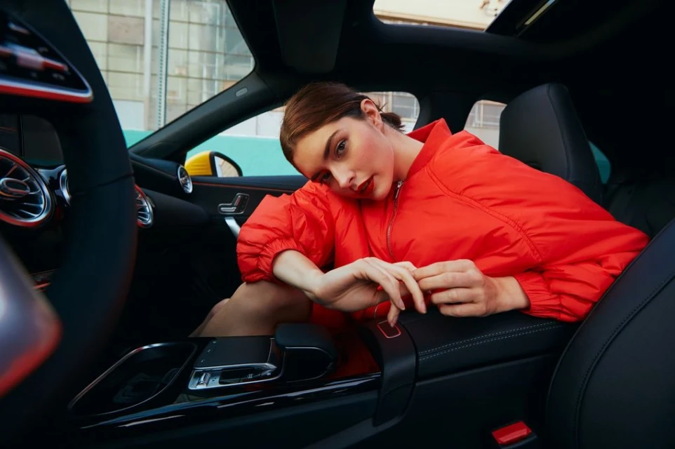 Woman in a red jacket poses in a car interior with black leather seats and a glimpse of an urban setting outside the car window.