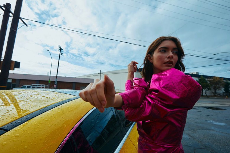 Woman in a pink satin dress stretches next to a yellow car on an urban street under a cloudy sky. Electric poles are visible.