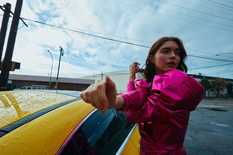 Woman in a pink satin dress stretches next to a yellow car on an urban street under a cloudy sky. Electric poles are visible.