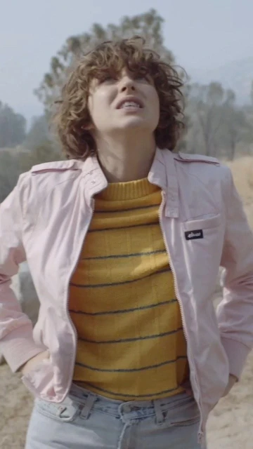 Woman in a pink jacket and yellow striped shirt stands outdoors in a sunlit, grassy environment with hazy, distant trees in the background.