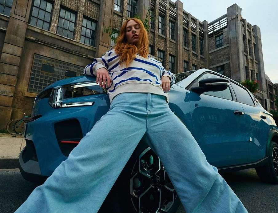 Red-haired woman in casual outfit leaning on a blue SUV against a backdrop of an urban brick building and city street.