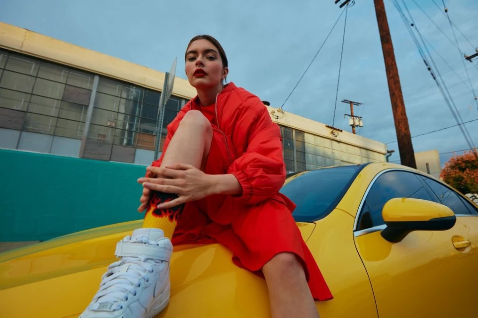 A young woman in a red jacket and skirt sits on the hood of a yellow sports car in an urban setting, wearing white high-top sneakers and flame socks.