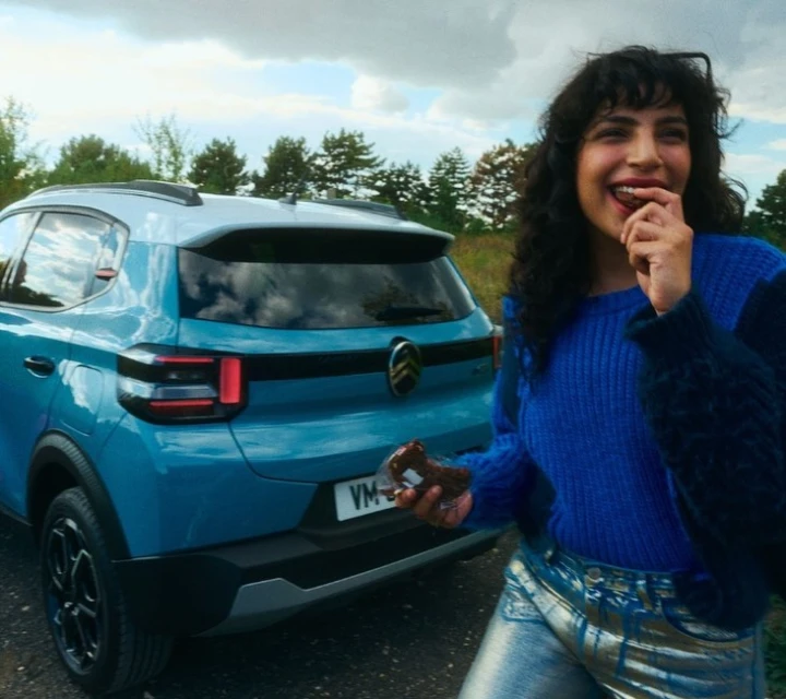 Woman in blue sweater eats chocolate treat near blue Citroen car in a rural outdoor environment, under a cloudy sky, portrait orientation.