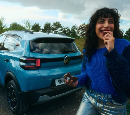 Woman in blue sweater eats chocolate treat near blue Citroen car in a rural outdoor environment, under a cloudy sky, portrait orientation.