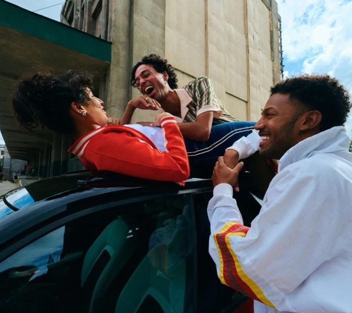 Three young adults, two males and one female, laugh on the roof of a black car in an urban setting with tall buildings in the background.