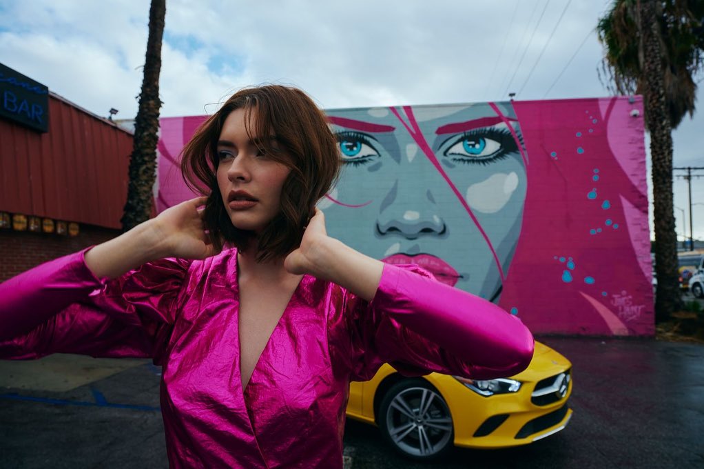A woman in a pink metallic dress poses outdoors with a car and mural art backdrop under a cloudy sky, creating a vibrant and stylish composition.