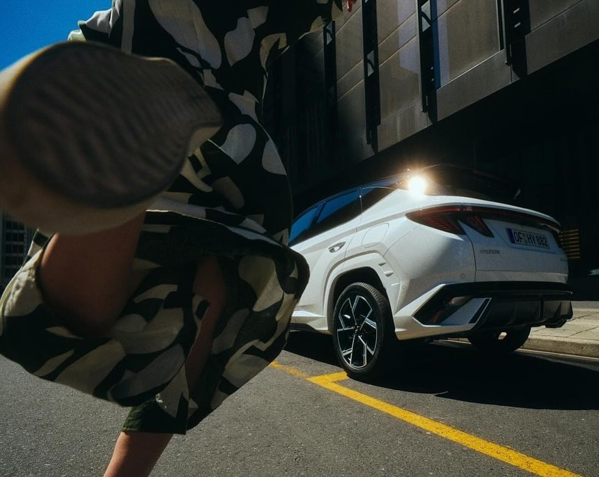 White SUV car photographed from the rear on asphalt road with blurred woman's legs. Building in the background and golden light.